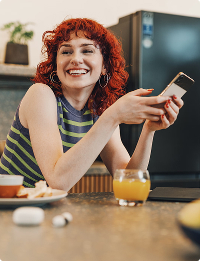 Ready to start A person with curly red hair sits at a kitchen counter, smiling while holding a smartphone, with a glass of orange juice and food on the table.