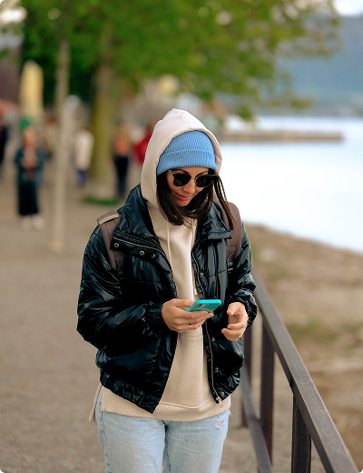 <span>No annual fee</span> for Standard plan A person wearing a blue beanie, sunglasses, a black jacket, and a beige hoodie walks outside by water while looking at a smartphone.