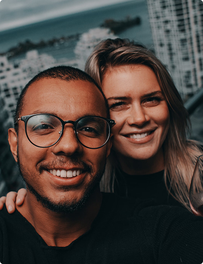Travel <span>perks</span> and <span>insurance</span> A man and a woman smiling for a selfie outdoors, with tall buildings and water visible in the background.