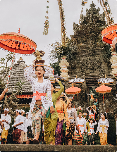 A group of people in traditional Balinese attire participate in a cultural ceremony in Bali, with women carrying offerings and ornate umbrellas, gathered in front of a decorated temple.