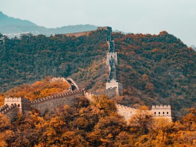 <span>Travel</span> card in <span>China</span> The Great Wall of China stretches across forested hills with autumn foliage, under a cloudy sky, showcasing the beauty and history of China.