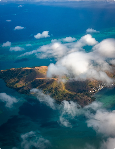 Aerial view of a Fiji island surrounded by blue-green water, with low clouds partially covering the land and coastline.