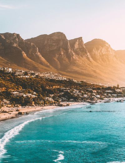 A scenic South African coastline with turquoise water, gentle waves, sandy beach, coastal town, and rugged mountains under a warm, golden sunset.