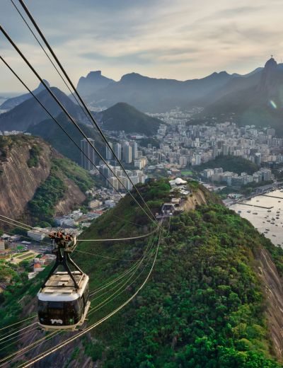 A cable car in South America travels above a green hillside toward a city with mountains in the background and water to the right, under a cloudy sky.