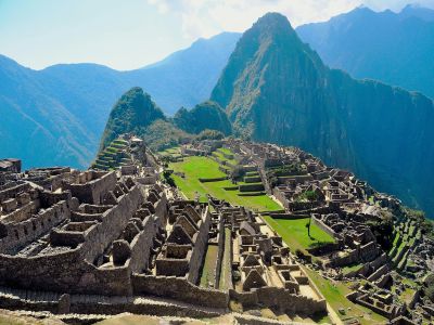 Ruins of Machu Picchu, an ancient Incan city in South America, sit atop a mountain with green terraces and surrounding peaks under a clear sky.