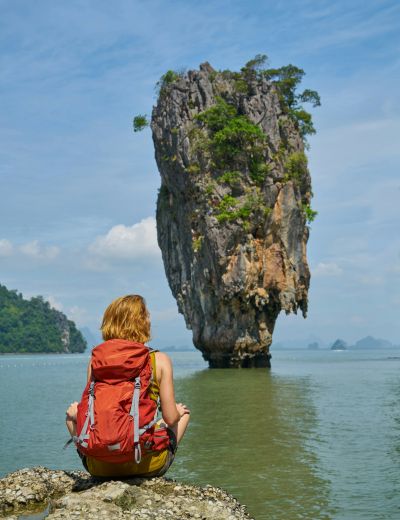 A person with a red backpack sits on a rock facing a tall limestone island rising from the water under Thailand’s blue sky.