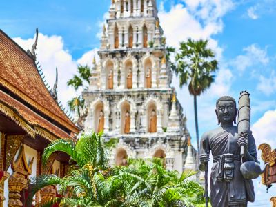 A stone Buddha statue stands in front of an ornate white temple with multiple tiers in Thailand, surrounded by palm trees and lush greenery under a blue sky.