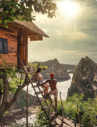 A couple stands on wooden steps leading to a treehouse, overlooking a scenic coastal landscape with rocky cliffs and sunlight streaming through clouds.