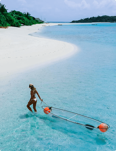 A woman in a white bikini pulls a transparent kayak with orange floats through shallow, clear blue water along a tropical beach.