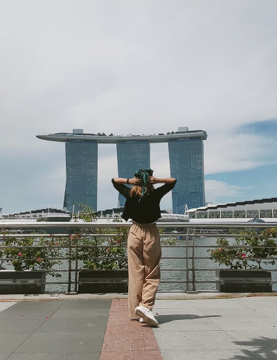 A person stands facing the Marina Bay Sands hotel in Singapore, with hands on their head, near a waterfront railing under a partly cloudy sky.