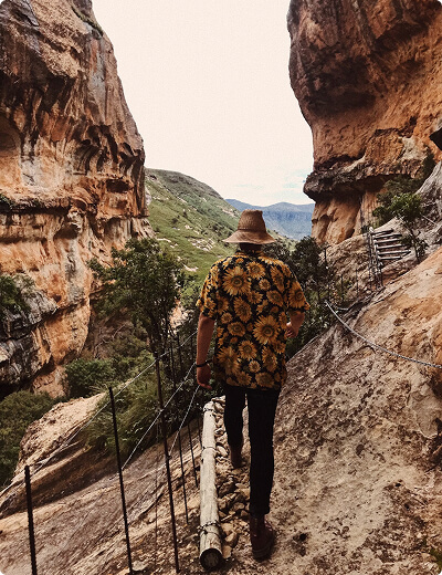 Person wearing a sunflower shirt and hat walks along a narrow rocky path between cliffs, with green hills visible in the background.