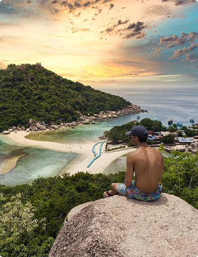 A man in swim trunks and a cap sits on a rock overlooking a tropical island, clear water, and a sandy beach at sunset.