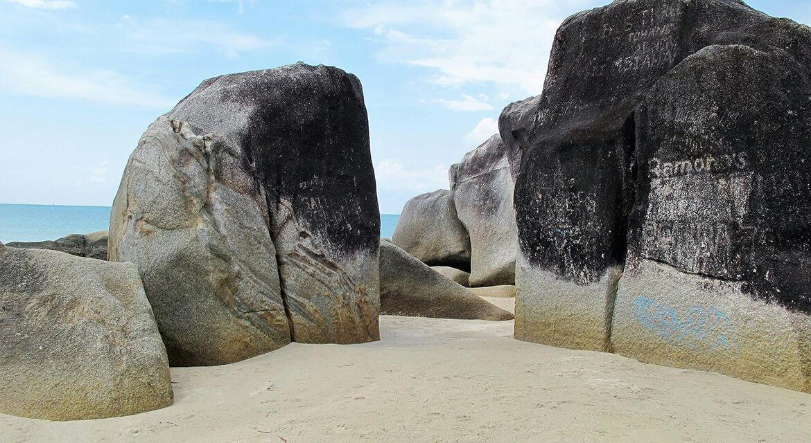 A beach scene featuring large, weathered granite rock formations with striking color contrast—light tan at the base and darker hues above, possibly from mineral deposits or erosion.