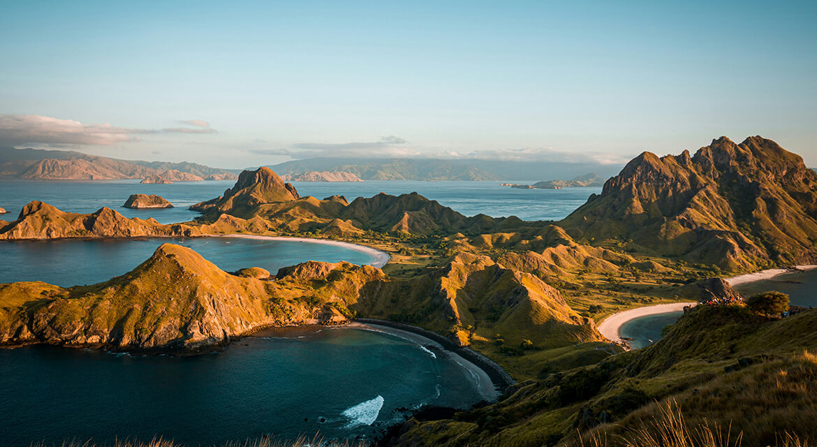 Panoramic view of Padar Island with sharp green hills, crescent-shaped beaches, and turquoise waters under golden sunlight.