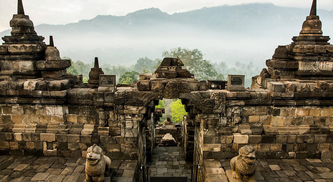 Stone archway and stupas of an ancient temple complex with misty mountains in the background.
