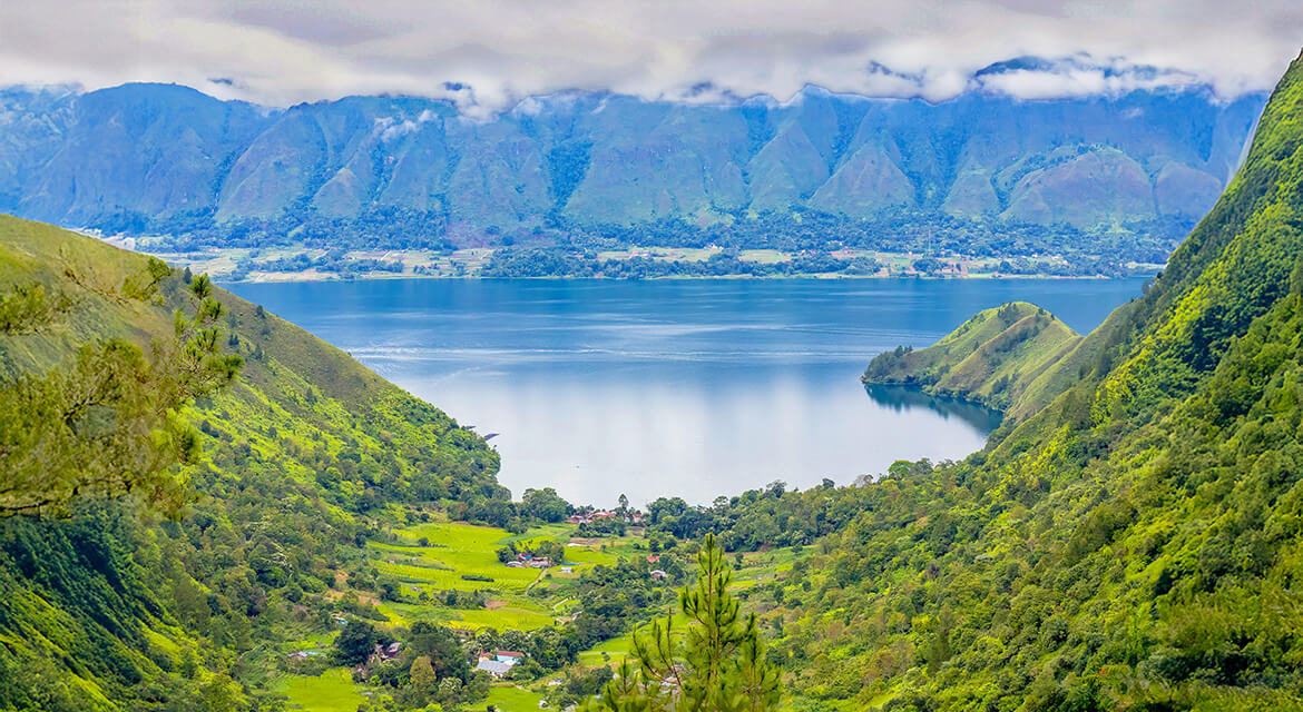 A panoramic view of a tranquil lake nestled in a lush green valley, surrounded by steep forested hills and towering mountains partially veiled by clouds.