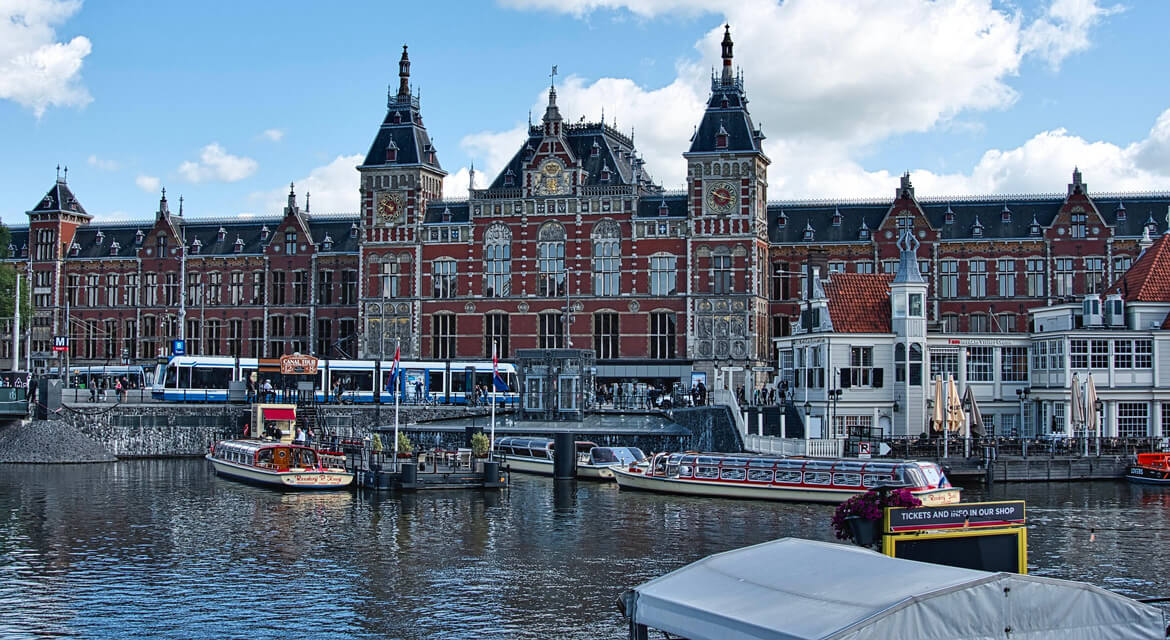 View of Amsterdam Centraal Station with Gothic-Renaissance architecture, boats docked in the canal, and trams on the street, capturing the city’s blend of historic elegance and modern transport convenience