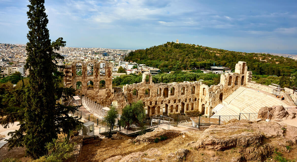 View of the Odeon of Herodes Atticus with tiered stone seating and arched stage facade, set against the cityscape of Athens and distant hills, reflecting Greece’s rich historical and cultural heritage