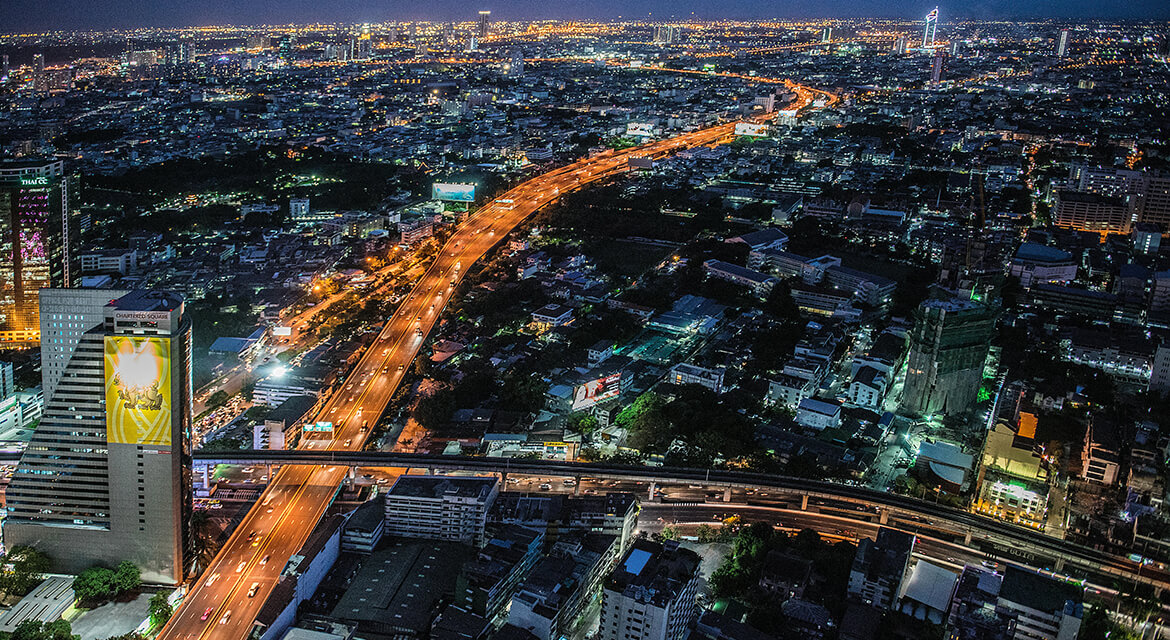 Nighttime aerial view of Bangkok with illuminated highways and dense urban buildings, capturing the city’s dynamic energy and blend of tradition and modernity