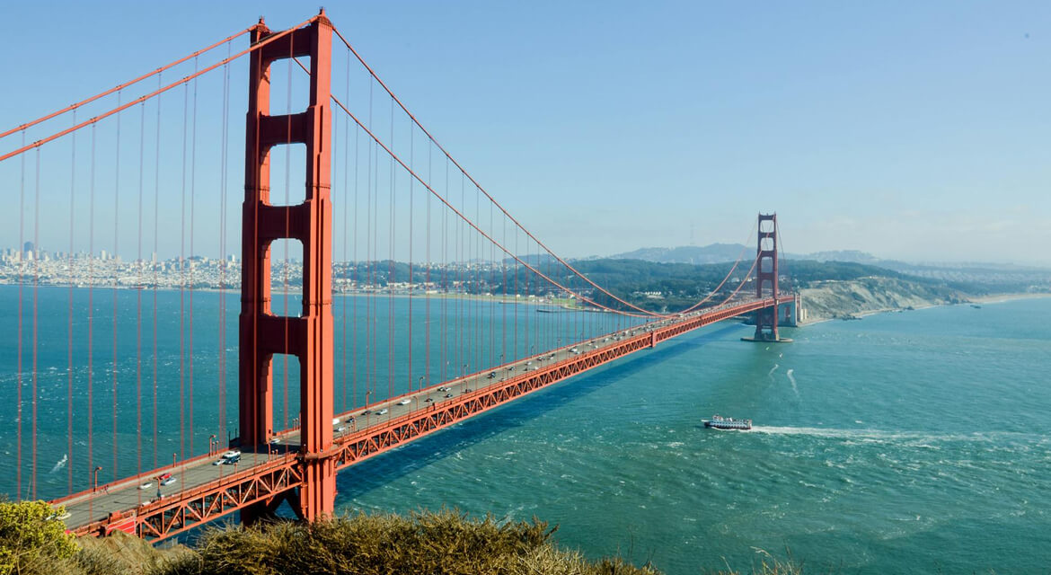The Golden Gate Bridge stretching across the San Francisco Bay under a clear blue sky, with vehicles crossing and a boat below, symbolizing the city’s iconic engineering, scenic beauty, and adventurous spirit.