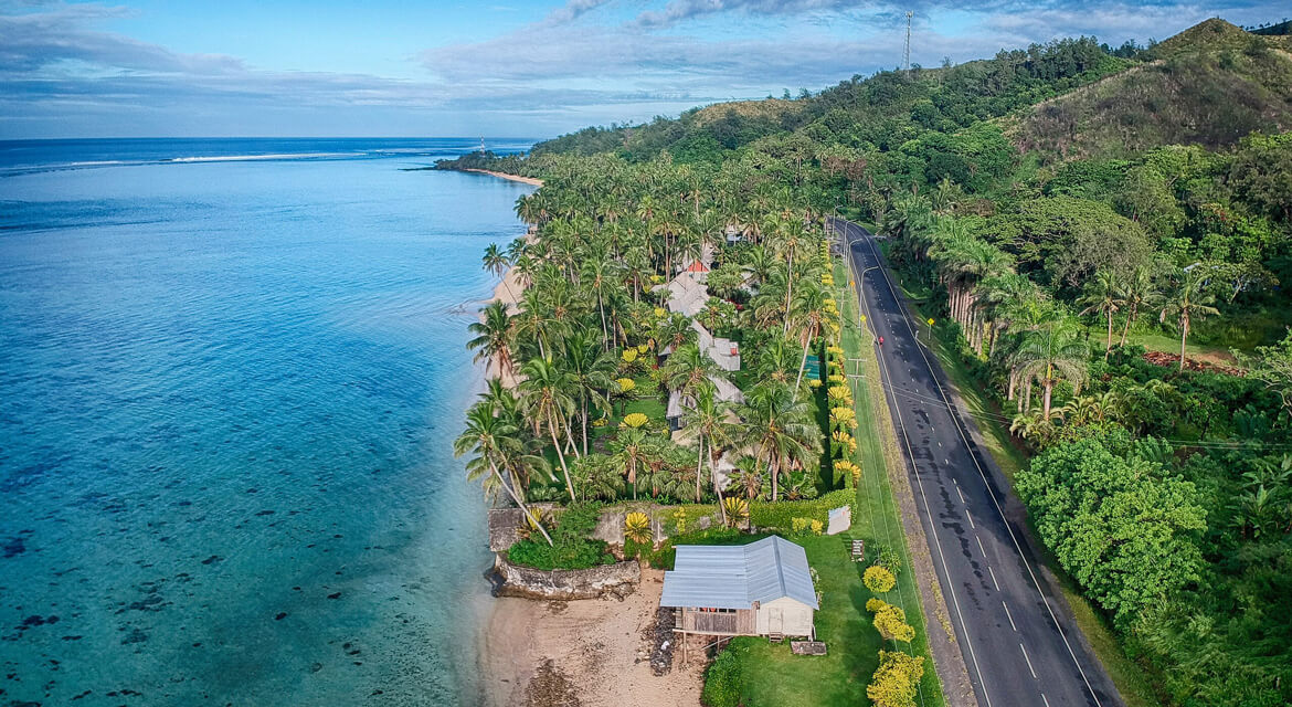 Aerial view of a winding coastal road beside clear blue ocean, sandy beach, and tropical greenery with cottages nestled among palm trees.