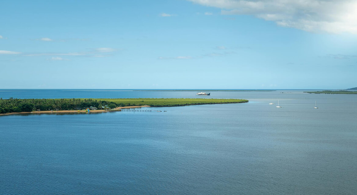 Wide coastal scene with calm bay waters, a narrow green strip of land, a pier, and distant ships under a clear sky, reflecting Suva’s blend of nature and activity.