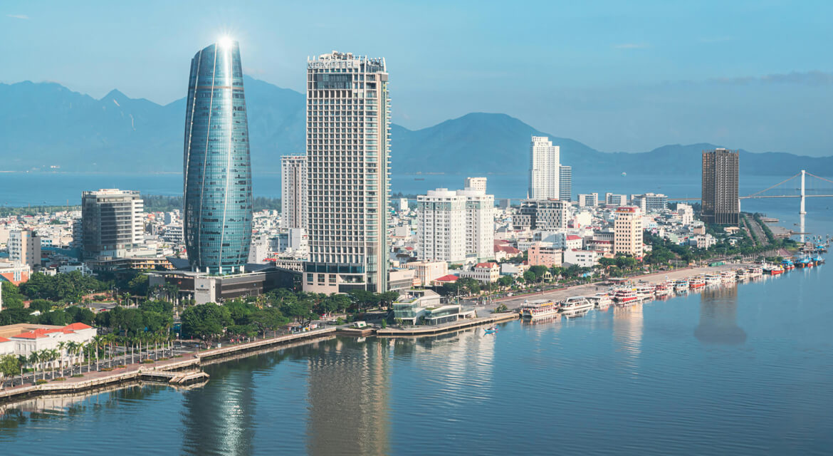 Panoramic daytime view of Da Nang cityscape with modern glass skyscrapers, calm waterfront, and distant mountains, showcasing the city’s blend of natural beauty and contemporary architecture