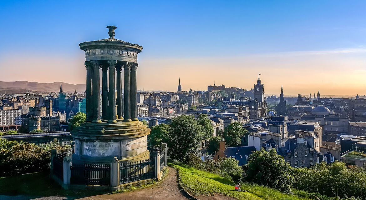 Panoramic view from Calton Hill featuring the Dugald Stewart Monument in the foreground and Edinburgh Castle with historic cityscape in the warm glow of sunset