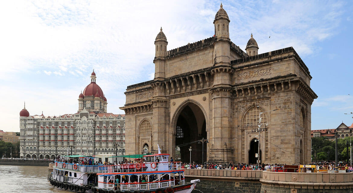 The Gateway of India arch and Taj Mahal Palace Hotel in Mumbai with boats docked at the waterfront and crowds enjoying the lively atmosphere