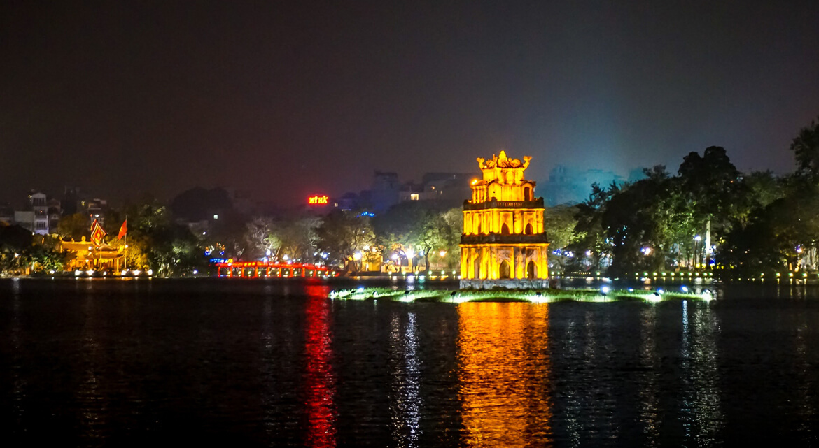 Nighttime view of Hoan Kiem Lake in Hanoi with illuminated Turtle Tower and red Huc Bridge reflecting on the water, surrounded by trees and city lights showcasing the city’s historic and vibrant atmosphere