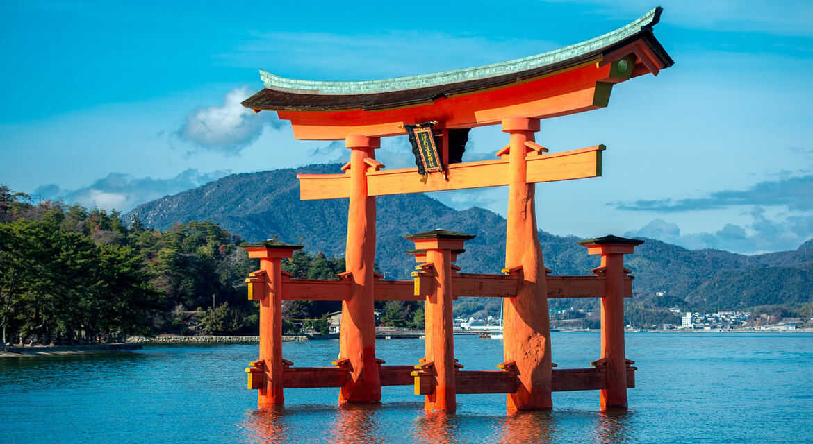Floating torii gate of Itsukushima Shrine in Hiroshima standing in calm waters with forested hills and blue sky in the background, symbolizing the spiritual and architectural elegance of Japan