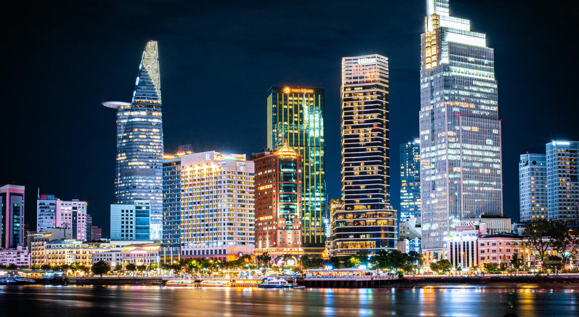 Nighttime cityscape of Ho Chi Minh City with illuminated skyscrapers including Bitexco Financial Tower reflecting on the river, showcasing the city’s dynamic urban growth and vibrant atmosphere