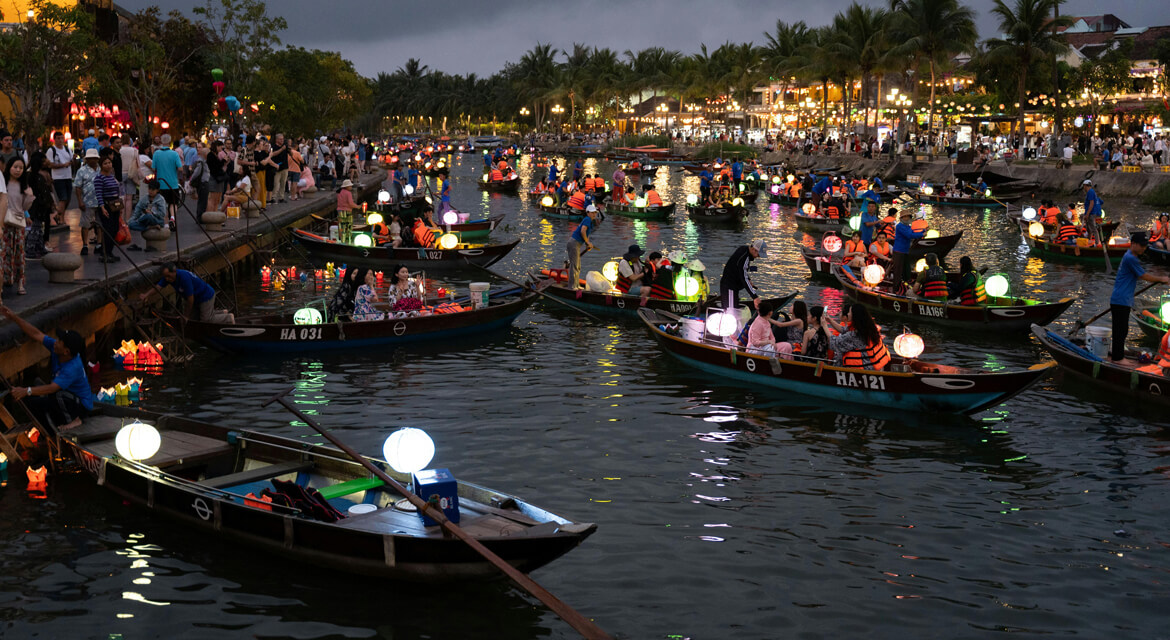 Festive river scene in Hoi An with boats adorned in colorful lanterns and crowds on the banks celebrating at dusk, capturing the vibrant spirit of Vietnamese cultural traditions