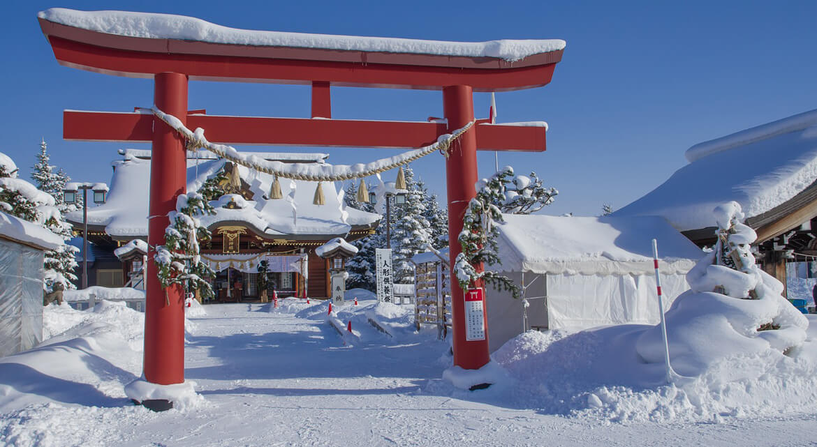 Snow-covered Shinto shrine in Hokkaido with a red torii gate and traditional architecture surrounded by trees under a clear blue sky, capturing the serene beauty of Japan’s winter landscape