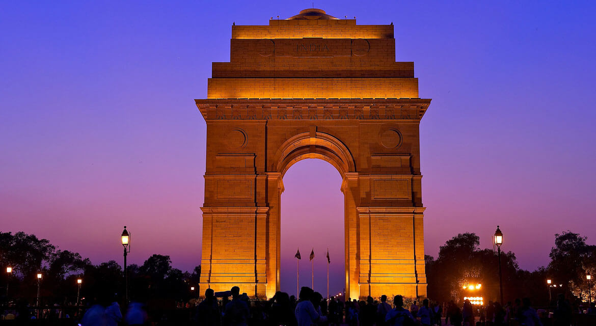 India Gate glowing under warm lights against a twilight sky in New Delhi with silhouetted trees and visitors in the foreground