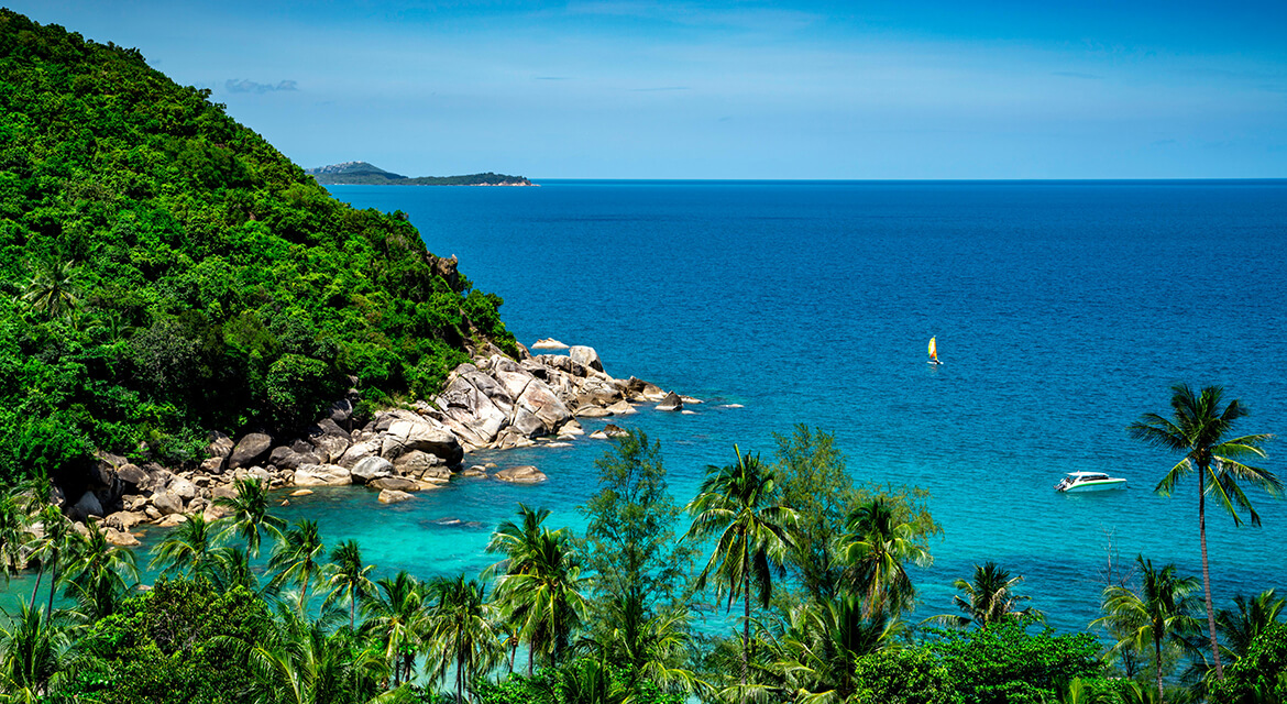 Coastal view of Koh Samui with turquoise waters, palm trees, a motorboat, and windsurfer near a forested shoreline under a clear blue sky, capturing the island’s serene atmosphere and recreational charm
