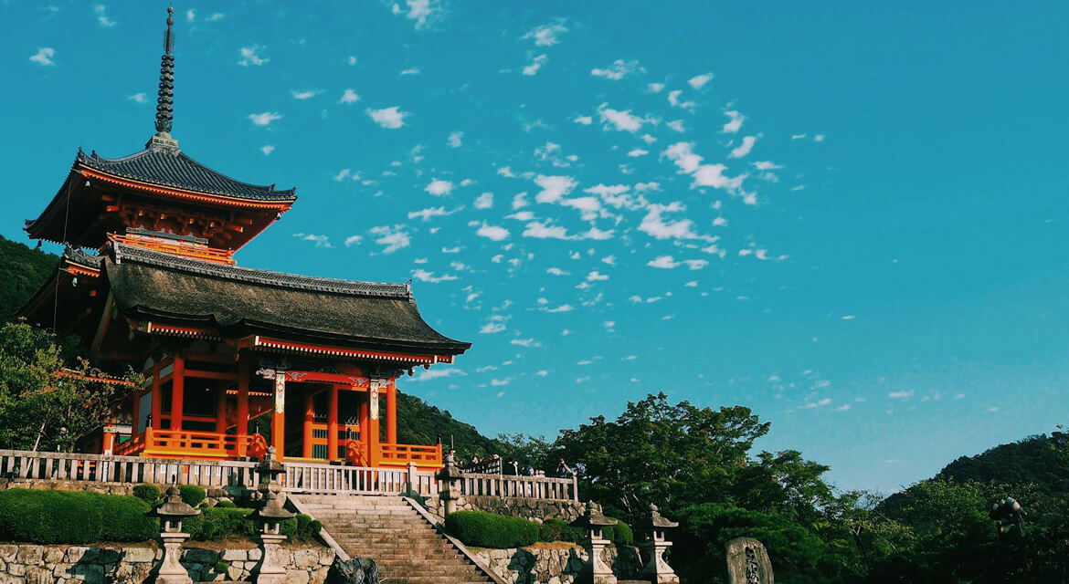 Traditional Japanese temple in Kyoto with a curved roof and vibrant red-orange structure surrounded by stone lanterns and lush greenery under a clear sky