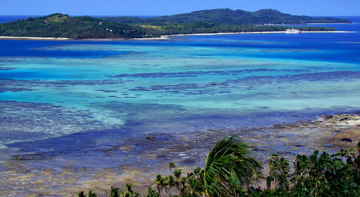 Tropical coastline with palm trees, turquoise shallows, coral reefs, and a distant island with white sand and a boat anchored nearby.
