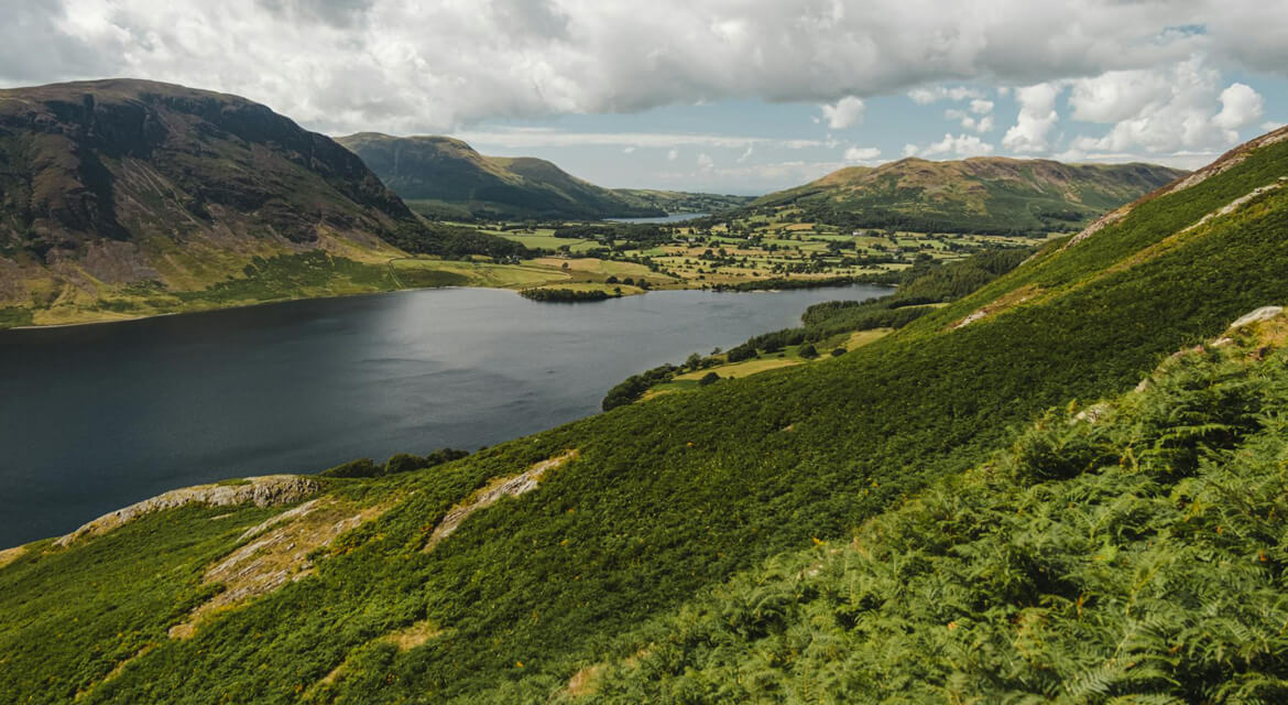 Tranquil lake nestled in a green valley surrounded by rolling hills and dense vegetation under a partly cloudy sky in the Lake District England