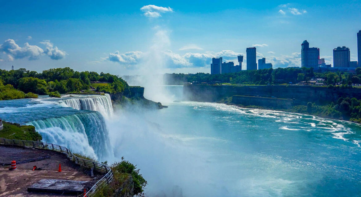 View of Niagara Falls with water crashing over the edge, mist rising into the air, and city skyline in the background across the river