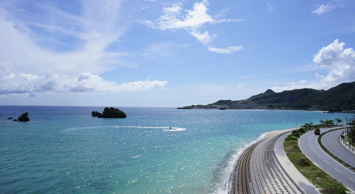 Scenic coastal view of Okinawa with turquoise ocean, curving shoreline road, rocky islets, and a jet ski trail under a clear blue sky, capturing the island’s tropical charm and natural beauty