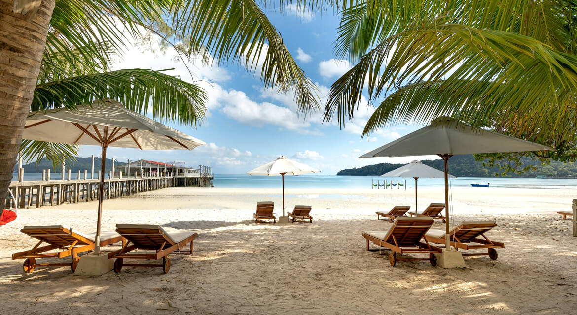 Serene beach scene in Phuket with lounge chairs under white umbrellas, palm trees, a wooden pier over turquoise waters, and swings in the sea, capturing the island’s relaxing and playful coastal vibe
