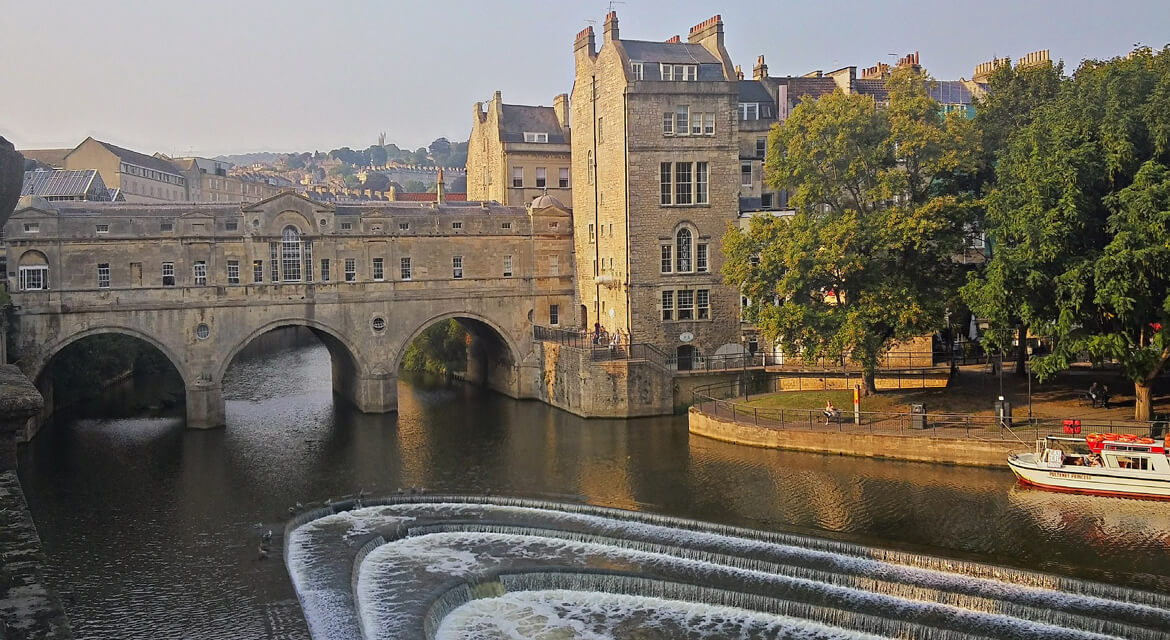 Historic view of Pulteney Bridge with shops lining both sides and a curved weir creating ripples on the River Avon surrounded by classical architecture in Bath England