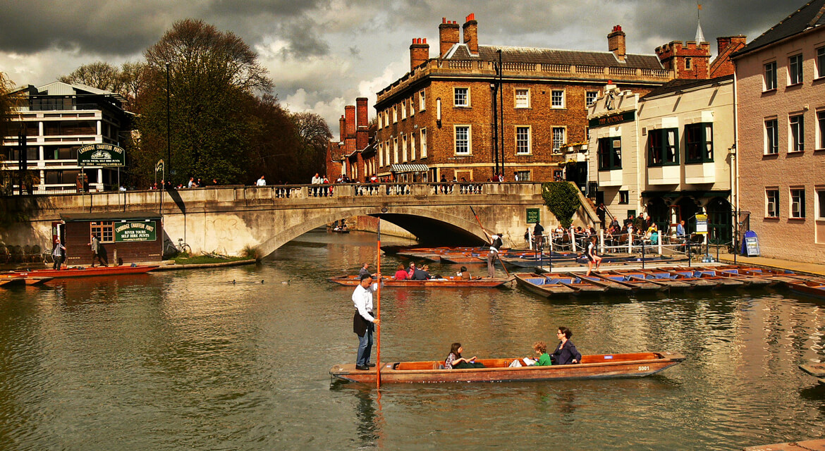 Scenic riverside view in Cambridge featuring people punting near a red-brick building and stone bridge with historic university charm and relaxed atmosphere