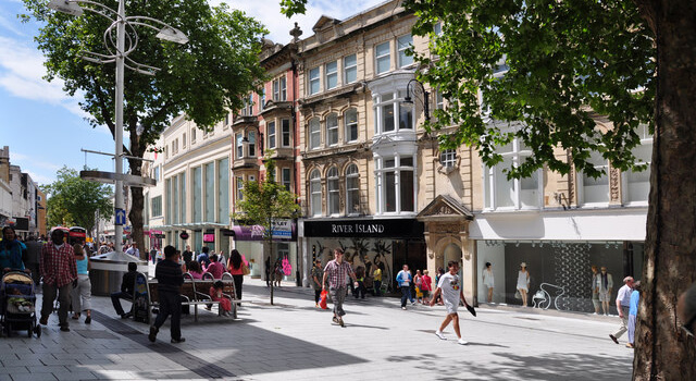 Lively urban scene on Queen Street in Cardiff featuring people walking and shopping among historic and modern buildings with tree-lined paths and retail storefronts