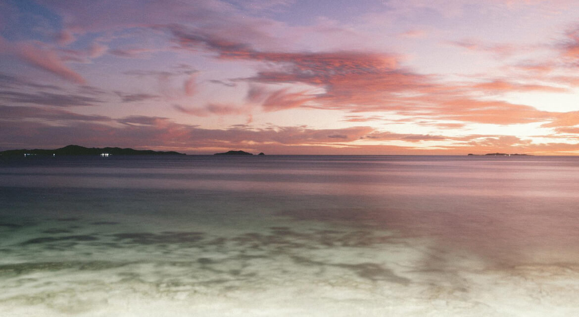 Serene beach scene at sunrise with soft waves, colorful sky in pink and orange hues, and distant islands silhouetted on the horizon.