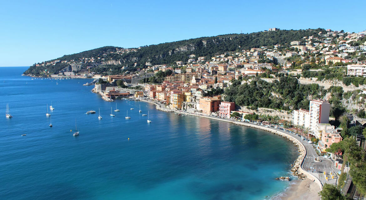 A vibrant coastal town nestled along a curved shoreline with clear blue waters. Colorful buildings with red-tiled roofs climb the hillside, surrounded by lush greenery.