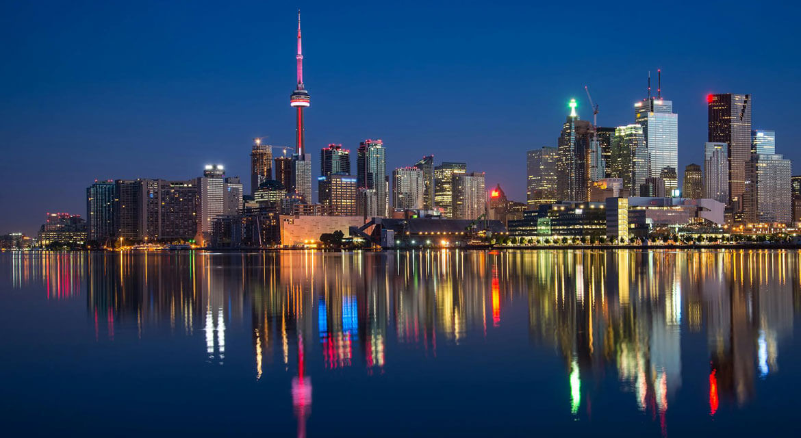Night view of Toronto’s cityscape with the CN Tower lit in red surrounded by high-rise buildings and colorful reflections on the calm waterfront